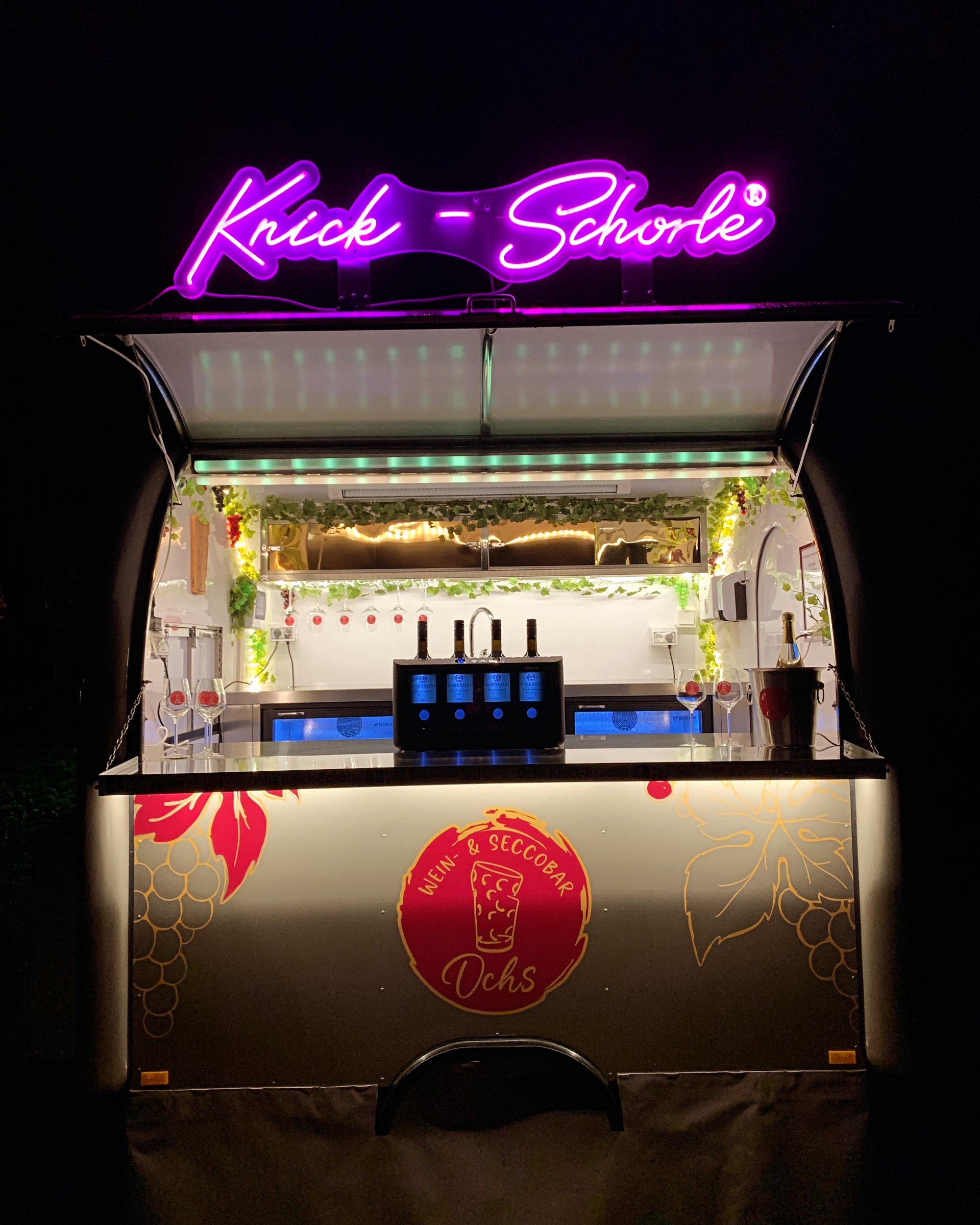 Seccobar Ochs Ein stilvoll beleuchteter Getränkewagen bei Nacht. Oben leuchtet ein pinkfarbenes Neonschild mit der Aufschrift „Knick-Schorle“. Der Wagen ist als Wein- und Seccobar gestaltet, dekoriert mit Weinranken und LED-Lichtern. Auf der Front prangt das Logo „Wein & Seccobar Ochs“ mit einem Glasmotiv.